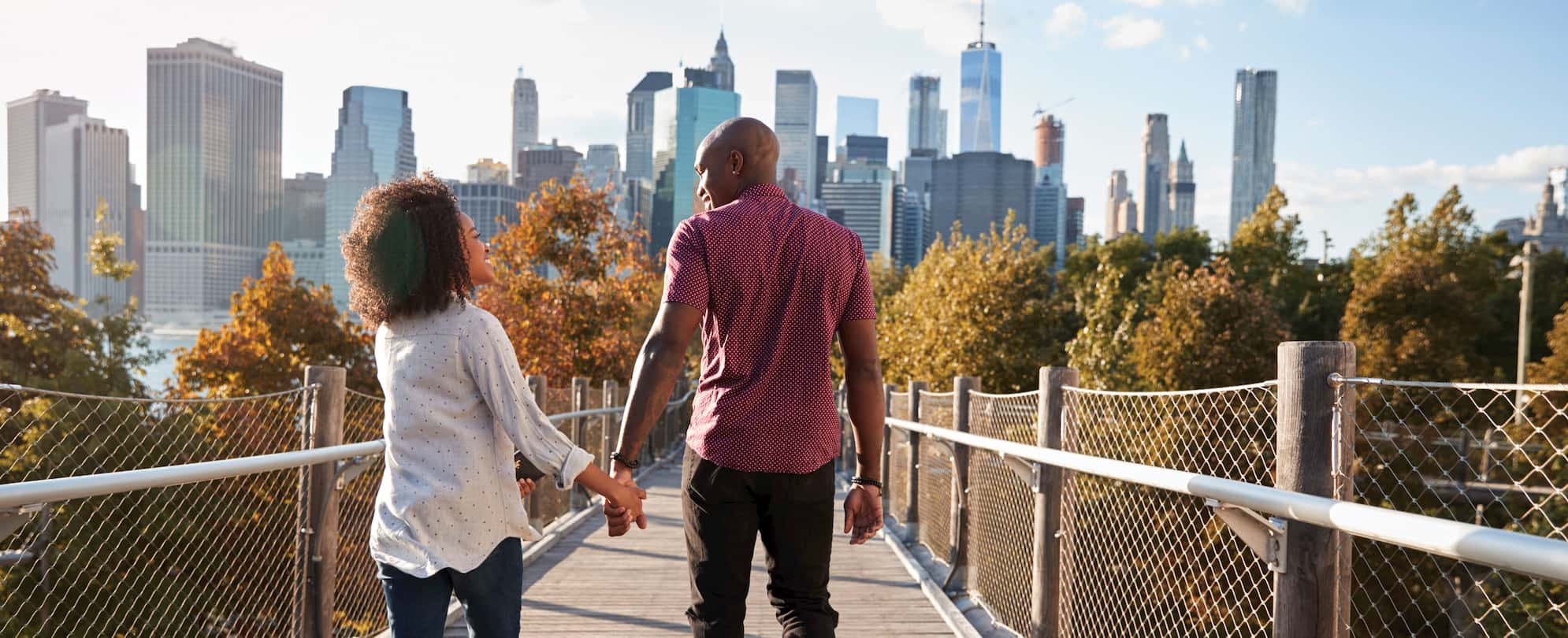 A man and a woman hold hands as they walk down a nature bridge toward the city