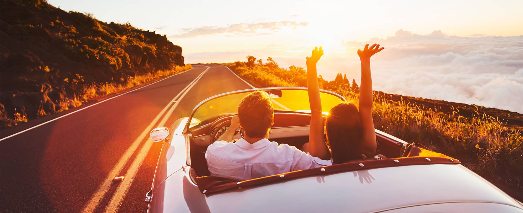 A man and a woman cheerfully drive off into the sunset in a convertible
