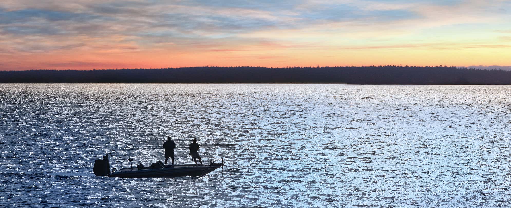 Two people stand on a small boat in the middle of a lake looking out over the sunset in Grand Lake, Oklahoma