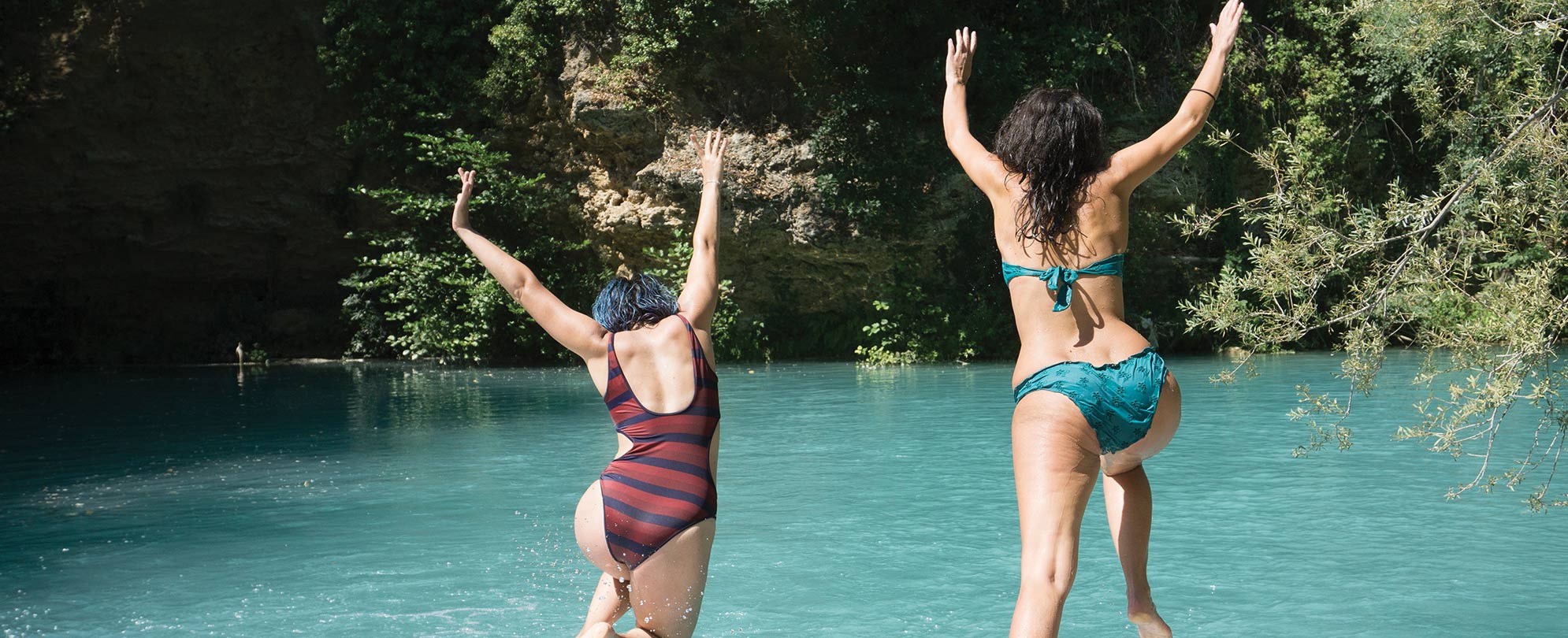 View from behind as two women in bathing suits jump with their arms in the air into a spring surrounded by cliffs and trees.
