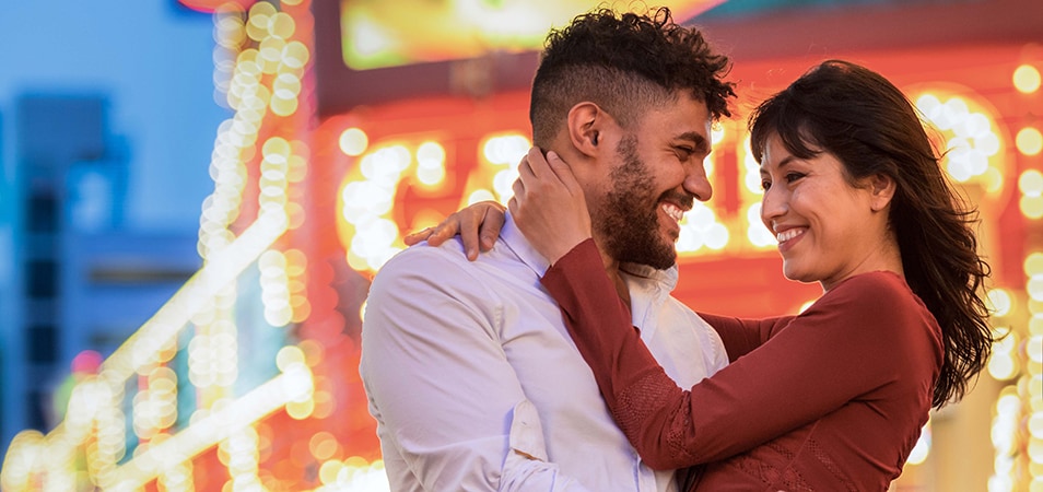 A smiling couple embraces and looks into each other's eyes with the bright lights of Las Vegas in the background.
